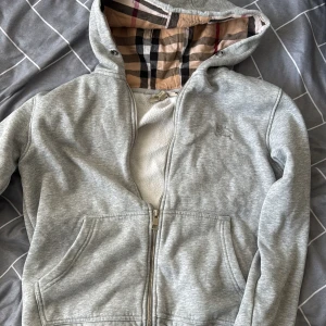Grå huvtröja från Burberry - Säljer en stilren grå huvtröja från Burberry med klassiskt rutigt foder i huvan. Tröjan har dragkedja framtill och två praktiska fickor. Perfekt för en avslappnad look med en touch av lyx. Storlek S, pris kan diskuteras och byten kan tas emot ifall det är i samma prisklass. OBS! Dragkedjan är slö men kan fixas billigt hos en bra skräddare. 