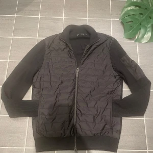 J.Lindberg jacka - Snygg svart jacka från J.Lindeberg med quiltad framsida och ribbade ärmar. Jackan har dragkedja och en stilren design som passar perfekt för höst och vår. Den är tillverkad i en kombination av material som ger både värme och stil.