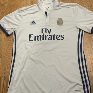 Vit Real Madrid fotbollströja från Adidas - Säljer en vit Real Madrid fotbollströja från Adidas med korta ärmar och Climacool-teknologi. Tröjan har klubbens emblem och sponsorlogga på framsidan samt 'Sergio Ramos' och nummer 4 på baksidan. La Liga-märke på ärmen.