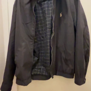 Svart jacka från Polo Ralph Lauren - Snygg svart jacka från Polo Ralph Lauren med ett stilrent rutigt mönster på insidan. Jackan har en dragkedja framtill och en liten broderad logga på bröstet. Perfekt för en klassisk look. Nypris: 2800 