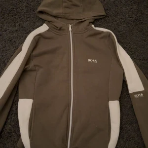 Brun huvtröja från Boss - Säljer en brun huvtröja från Boss med dragkedja och vita detaljer längs ärmarna. Perfekt för en avslappnad stil. Tröjan har en huva och är långärmad.