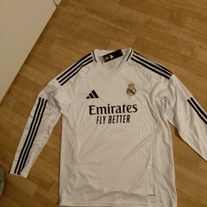 Vit långärmad Real Madrid fotbollströja från Adidas - Snygg vit långärmad fotbollströja från Adidas med Real Madrids emblem på bröstet. Tröjan har svarta ränder på ärmarna och texten 'Emirates Fly Better' på framsidan. Perfekt för alla Real Madrid-fans!