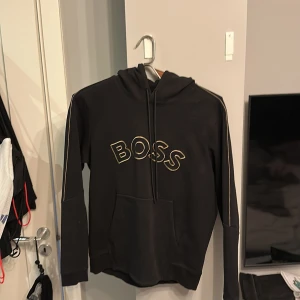 Svart hoodie från BOSS - Snygg svart hoodie från BOSS med stor logga på bröstet. Tröjan har en klassisk känguruficka och justerbar huva med dragsko. Perfekt för en stilren och bekväm look.