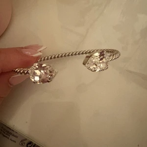 Silverfärgat armband med kristaller - Elegant armband i silverfärg med två stora glittrande kristaller. Armbandet har en vriden design som ger en unik touch. Perfekt för att lägga till lite glamour till din outfit. Köpt för 795 kr knappt använt då den är för stor för min arm.