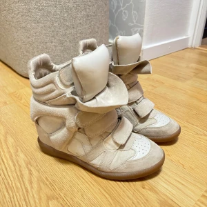Isabel Marant skor - Snygga beige sneakers med kilklack och kardborreband. Skorna har en unik design med vadderad plös och häl, vilket ger extra komfort. Perfekta för en trendig look.
