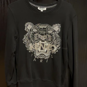 Svart sweatshirt från Kenzo med tigerbroderi - Snygg svart sweatshirt från Kenzo med ett ikoniskt tigerbroderi i silver på framsidan. Tröjan har rund hals och långa ärmar, perfekt för en stilren look. Passar bra till både jeans och kjol. 🐯✨