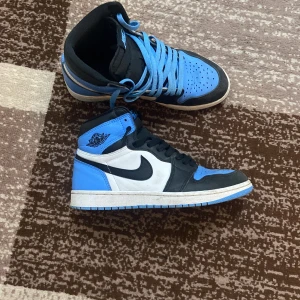 Nike Air Jordans  unc toe - Snygga Nike Air Jordans i blått, svart och vitt. Skorna har en klassisk design med snörning och en hög siluett. Perfekta för dig som vill ha en stilren och sportig look.