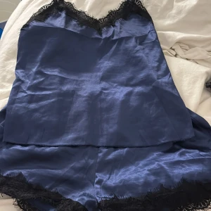 Marinblå linnetopp samt shorts med spetsdetaljer - Säljer en marinblå linnetopp samt shorts i satinliknande material med tunna axelband och v-ringning dekorerad med svart spets. Toppen har en glansig finish och justerbara band för perfekt passform. Perfekt för dig som gillar en chic och elegant look. Detta sett är perfekt för en mer chill hemma look