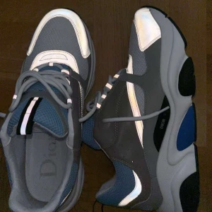 Dior B22 - Säljer ett par Dior b22 sneakers med cool design i blått, grått, vitt och svarta detaljer. Skorna har mesh och skinnpaneler, bred sula och reflekterande partier. Snygga snören och klassisk Dior-logga på sidan. Perfekta för dig som gillar statement sneakers. Passar storlek 45 möts i Sthlm