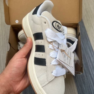 Adidas Campus i beige mocka - Snygga Adidas Campus sneakers i beige mocka med svarta läderdetaljer och klassiska tre ränder på sidan. Skorna har vit sula, breda vita skosnören och svart Adidas-logga på häl och plös. Perfekta för dig som gillar retrovibbar och streetstyle.