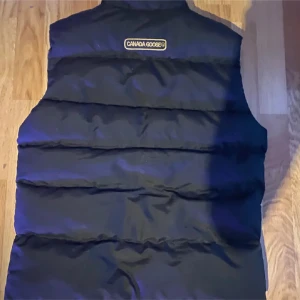 Svart dunväst från Canada Goose M - Svart dunväst från Canada Goose med klassisk broderad logga på bröstet och quiltad design. Västen har hög krage och dragkedja framtill. Perfekt för lager-på-lager och riktigt snygg till streetwear eller outdoor-stil. 