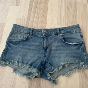 Blå jeansshorts med fransig kant - Snygga blå jeansshorts med låg midja och fransig, rå kant nertill. Klassisk femficksmodell med knappar på bakfickorna och slitna detaljer.💞💞