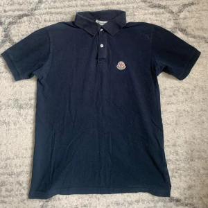 Mörkblå Moncler pikétröja - Snygg mörkblå pikétröja från Moncler med klassisk krage, två knappar och broderad logga på bröstet. Tillverkad i mjuk bomull och har korta ärmar. Perfekt för en clean och stilren look.