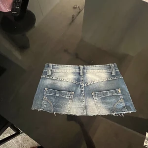 Blå jeans minikjol med rå kant - Supersnygg blå jeans minikjol med slitna detaljer och rå, fransig kant. Kjolen har fem knappar framtill, bälteshällor och klassiska fickor både fram och bak. Perfekt för dig som vill ha en edgy och trendig look.