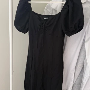 Svart kort puffärmad klänning - Svart klänning från Gina Tricot med puffärmar och söt knytning framtill. Modellen är kort och har en feminin, figurnära passform. Perfekt för dig som gillar en stilren look med trendiga detaljer.