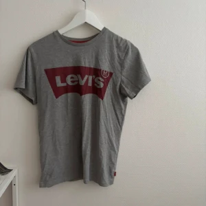 Grå Levi's t-shirt med röd logga - Snygg grå t-shirt från Levi's med klassisk röd logga framtill. T-shirten har rund halsringning och korta ärmar. Tillverkad i mjuk bomull som är skön att bära hela dagen. Perfekt för en avslappnad och stilren look.