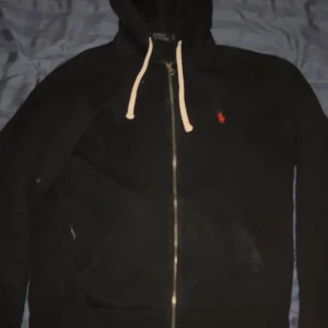 Svart hoodie från Polo Ralph Lauren - Svart hoodie från Polo Ralph Lauren med dragkedja och vit snörning i huvan. Klassisk röd logga broderad på bröstet. Mjuk och skön, perfekt för lager-på-lager. Långa ärmar och känguruficka framtill.