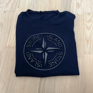 Mörkblå Stone Island t-shirt med tryck - Snygg mörkblå t-shirt från Stone Island med stort ikoniskt kompass-tryck på bröstet. Klassisk rund hals och korta ärmar. Tillverkad i mjuk bomull som känns skön mot huden. Perfekt för dig som gillar streetwear och vill sticka ut med en fet logga.