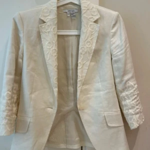 Broderad vit kavaj från Zara Basic - Snygg vit kavaj från Zara Basic med broderade blommor på krage och ärmar. Kavajen har klassisk passform, två fickor fram och är tillverkad i ett linne-liknande material. Perfekt för dig som vill sticka ut med stilrena detaljer och fräsch look.