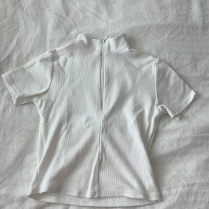 Vit ribbad polotopp med dragkedja - Vit ribbad polotopp med korta ärmar och dragkedja bak som kan öppnas hela vägen upp till kragen. Snygg och enkel design som passar till många olika outfits. Mjuk och stretchig känsla, perfekt för dig som gillar stilrena plagg med en twist.