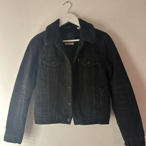 Svart jeansjacka med teddykrage Levi's - Svart jeansjacka från Levi's med mysig teddykrage och foder. Klassisk modell med bröstfickor, knappar framtill och detaljer i svart. Perfekt för dig som vill ha en cool och tidlös look med extra värme under höst och vinter. Storlek s