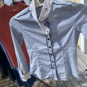 Randig ridinspirerad skjorta från CRW - Snygg blå- och vitrandig skjorta från CRW Classic Riding Wear. Skjortan har lång ärm, vita kontrastdetaljer på krage och manschetter samt mörkblå knappar och piping. Perfekt för dig som gillar ridstil och vill ha en klassisk look. ( rätt så liten runt axlarna )