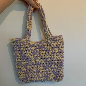 Virkad tote bag i lila och gult - Superfin virkad tote-bag i lila och gult med grovt mönster. Väskan har två rejäla handtag och är perfekt för att bära med sig allt man behöver. Den är mjuk och har en unik, handgjord känsla som verkligen sticker ut.