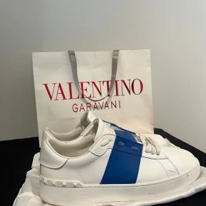 Valentino Garavani Open sneakers vit/blå - Snygga Valentino Garavani Open sneakers i vitt skinn med en bred blå rand över mitten. Klassisk chunky sula och coola nitar baktill. Skorna har rund tå, vita snören och en stilren design som sticker ut. Kvitto finns