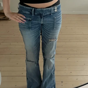 Blå bootcut jeans med slitningar - Snygga blå bootcut jeans med låg midja och slitningar vid knäna. Jeansen har dubbla knappar i midjan, stora framfickor och klassiska bakfickor. Perfekt för dig som gillar en avslappnad och trendig look.