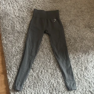 Grå seamless leggings från Gymshark - Mörkgrå seamless leggings från Gymshark med hög midja och diskret mönstrad struktur längs benen. Stretchigt material som sitter tight och formar sig efter kroppen. Gymshark-logga på höften och vid vristen. Byxorna har en skåra vid rumpan men det går att fixa och man syr ihop.