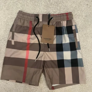 Rutig Burberry shorts med snörning - Snygga shorts från Burberry med klassiskt rutigt mönster i beige, svart, röd och blå. De har elastisk midja med svart snörning och sidofickor. Perfekta för sommaren när du vill sticka ut med en lyxig vibe.