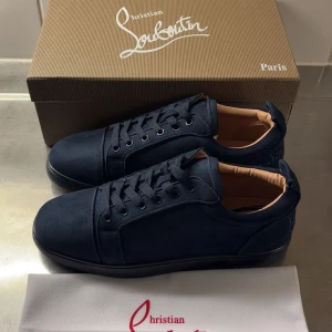 Mörkblå sneakers från Christian Louboutin - Snygga mörkblå sneakers från Christian Louboutin i mocka med klassisk röd sula. Skorna har rund tå, svarta snören och diskreta sömdetaljer. Insidan är ljusbeige och sulan har Louboutin-loggan i rött. Perfekta för dig som vill sticka ut med stil.