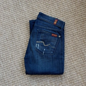Mörkblå jeans från 7 For All Mankind - Snygga mörkblå jeans från 7 For All Mankind med straight leg-modell. Klassisk femficksdesign, diskreta slitningar och dekorativa detaljer på bakfickorna. Jeansen är i mjukt stretchigt  denimtyg och har en normal passform som sitter skönt hela dagen. Storlek 25. Mycket välvårdade. Slitaget är i designen. 