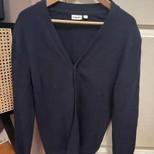 Mörkblå kofta från Kappahl - En stilren mörkblå kofta från Kappahl med v-ringning och lång ärm. Enkel och klassisk design som passar perfekt till lager-på-lager. Mjuk och skön, troligtvis i bomull eller stickad kvalitet. Perfekt för kyligare dagar.