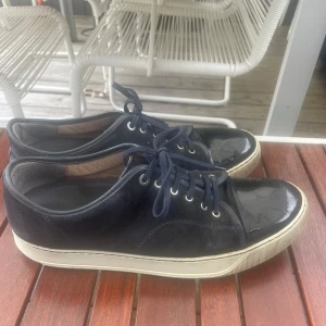 Lanvin cap toe  - Snygga svarta sneakers från Lanvin med ovandel i mocka och lackad tå. Skorna har vita platta sulor, svarta snören och silverfärgade öljetter. Perfekt för dig som gillar en stilren men ändå lyxig look.