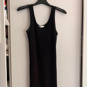 Svart ribbad långklänning - En svart, ribbad långklänning med ärmlös design och rund halsringning. Klänningen har en tight passform och är tillverkad i ett stretchigt material som sitter snyggt på kroppen. Perfekt för dig som gillar en clean och stilren look. Aldrig använd 