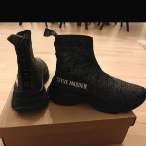 Svarta sockboots från Steve Madden - Helt nya!Trendiga svarta sockboots från Steve Madden med glittrig finish och tydlig logga på sidan. Skorna har en rund tå, platt sula och är tillverkade i stretchigt syntetmaterial som sitter tajt runt ankeln. 