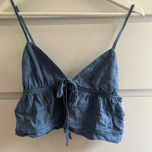 Blå linnetopp med knyt och spetsdetaljer - Supersöt blå linnetopp med smala axelband och knyt framtill. Toppen har volangiga partier och fina spetsdetaljer. Perfekt för varma dagar över en bikini. 