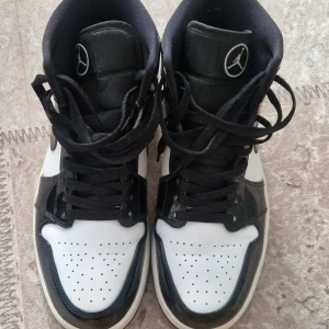 Nike Air Jordan 1 carbon fiber svartvita sneakers - Säljer ett par klassiska Nike Air Jordan 1 carbon fiber i svart och vitt. Skorna har Mid modell med snörning, svart Swoosh och Air Jordan-logga på sidan. Ovandelen är i skinn med perforerad tå och kontrasterande paneler. Perfekta för dig som gillar streetstyle och ikoniska sneakers.