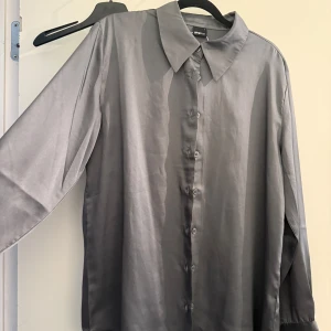 Grå satinliknande skjorta Gina Tricot - Stilren grå skjorta från Gina Tricot. Skjortan har lång ärm, klassisk krage och knäppning framtill. Materialet har en lätt glansig satinlook som ger en snygg touch till outfiten. Perfekt att styla med jeans eller kjol för en trendig vibe. Endast använd en gång. 