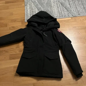 Svart dunjacka från Canada Goose - Säljer en svart dunjacka från Canada Goose med huva och klassisk logga på ärmen. Jackan har dragkedja, stora fickor framtill och en extra ficka på ärmen. Perfekt för kalla dagar och riktigt snygg streetstil.