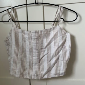 Randig linnetopp från Gina Tricot - Superfin randig topp i linne från Gina Tricot i beige och vitt. Toppen har smala axelband, croppad passform och smockad rygg för extra komfort. Har tidigare suttit knappar där fram som jag tagit bort då jag tyckte den var snyggare utan dom😊 vill bara nämna det för det kan synas om kollar man riktigt nära, men inget man tänker på💕 därför jag endast sätter den som bra skick, inga defekter annars 