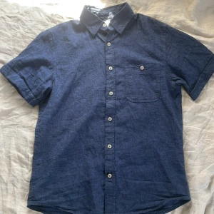 Blå linneskjorta från Dressmann - Snygg blå kortärmad skjorta från Dressmann i linnemix. Klassisk krage, vita knappar framtill och en bröstficka. Perfekt för varma dagar med sin lätta och luftiga känsla. Storlek S. Utmärkt skick då den knappt är använd. 