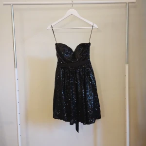 Svart glittrig kortklänning Model Behaviour - Supersnygg svart kortklänning från Model Behaviour, axelbandlös och hjärtformad urringning. Klänningen är täckt av glittriga paljetter och har ett satinband i midjan som knyts baktill. Perfekt för dig som vill sticka ut med en festlig look!