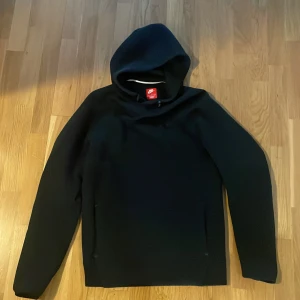 Svart nike tech fleece hoodie - Svart hoodie från Nike med diskret logga på bröstet. Tröjan har huva med snörning och långärmad design. Perfekt för dig som gillar en clean och sportig stil. Materialet känns mjukt och följsamt, och passformen är normal.