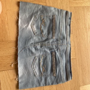Ljusblå jeanskjol med strassdetaljer - Säljer min ljusblå jeanskjol som jag har sytt själv och satt på snygga strassdetaljer på fickorna. Den har också broderade rosetter på bakfickorna. Kjolen är 38 cm rakt över och är 28 cm lång. Är en aning för tajt för mig och därför säljer jag den❤️