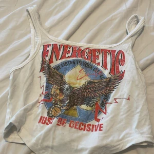 Vit croppad linne med tryck - Säljer en vit croppad linne med breda axelband och färgglatt tryck framtill med texten 'Energetic' och en stor örn. Toppen har röda, blåa och gula detaljer och är perfekt för dig som gillar statement-plagg.