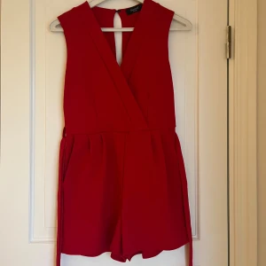 Röd omlott playsuit från Sisters Point - Supersnygg röd playsuit från Sisters Point med omlott framtill och knytband i midjan. Ärmlös modell med v-ringning och korta ben, perfekt för dig som vill sticka ut. Enkel men trendig design som passar till många tillfällen.