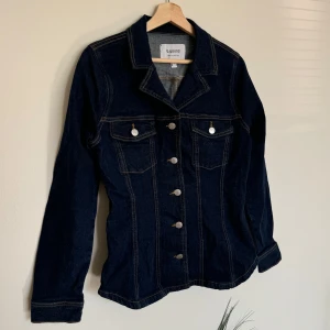 Mörkblå jeansjacka från b.young - Snygg mörkblå jeanströja med krage, silverfärgade knappar och två bröstfickor med lock. Jackan har kontrastsömmar och lång ärm. 
