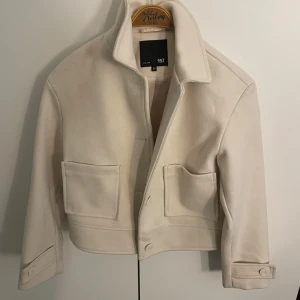 Vit ulljacka från 157, XS - Snygg vit jacka från 157 i ullmix med krage, stora fickor framtill och knappar vid ärmslut. Kortare modell som ger en clean och modern look. Perfekt för dig som gillar minimalistisk stil och vill ha något som sticker ut på ett stilrent sätt.